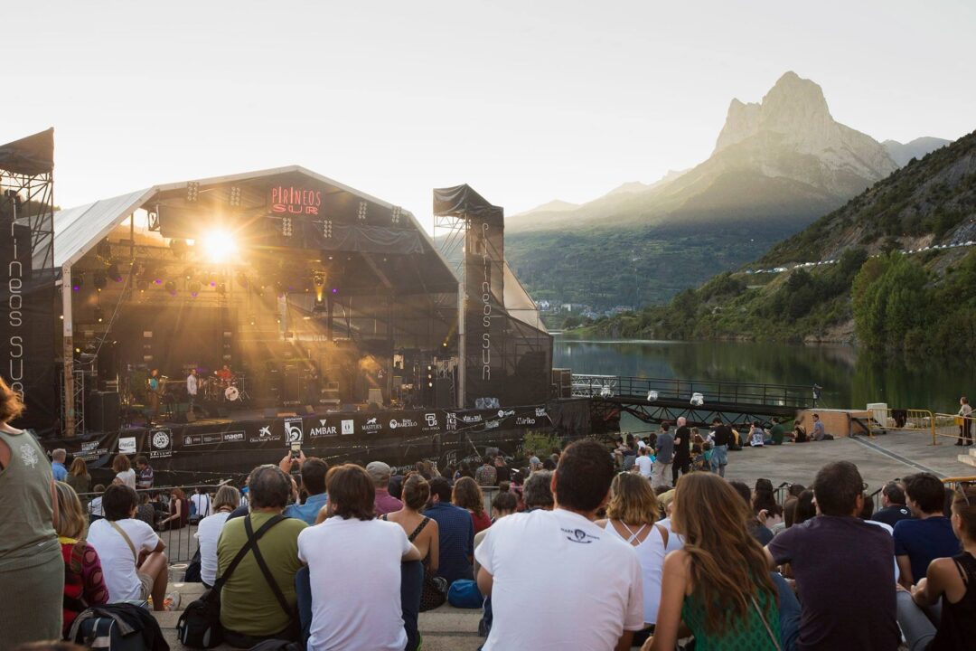 Público sentado frente al escenario flotante de Pirineos Sur con el pantano y las montañas al fondo.